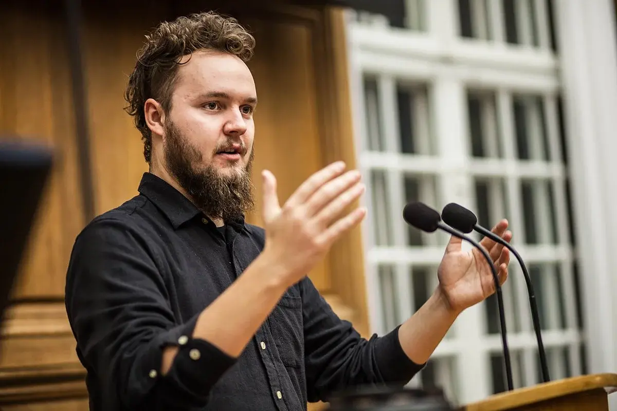 A man speaking at a podium with microphones in a parliamentary chamber.