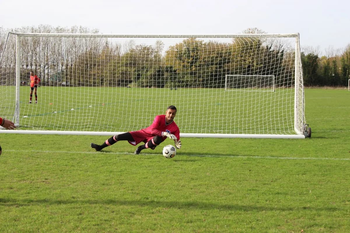 A goalkeeper dives to save a soccer ball near the goal on a grassy field.
