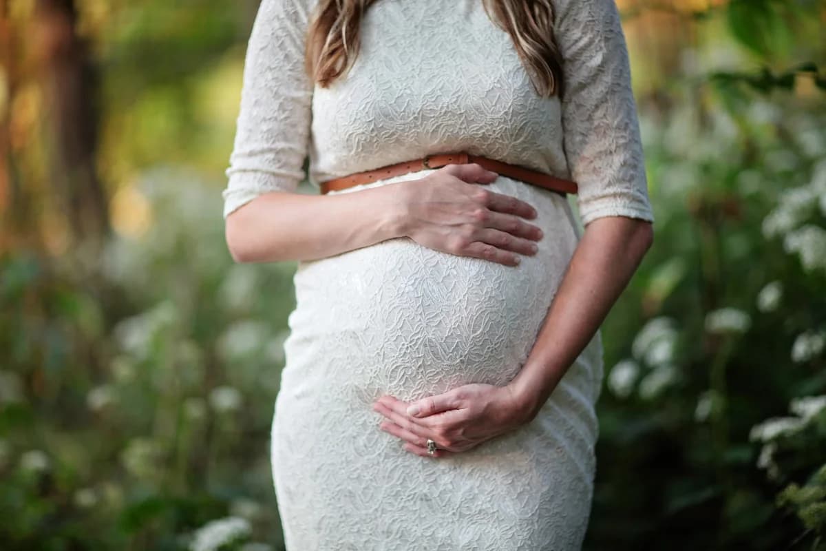 Pregnant woman in a white lace dress holding her belly outdoors.