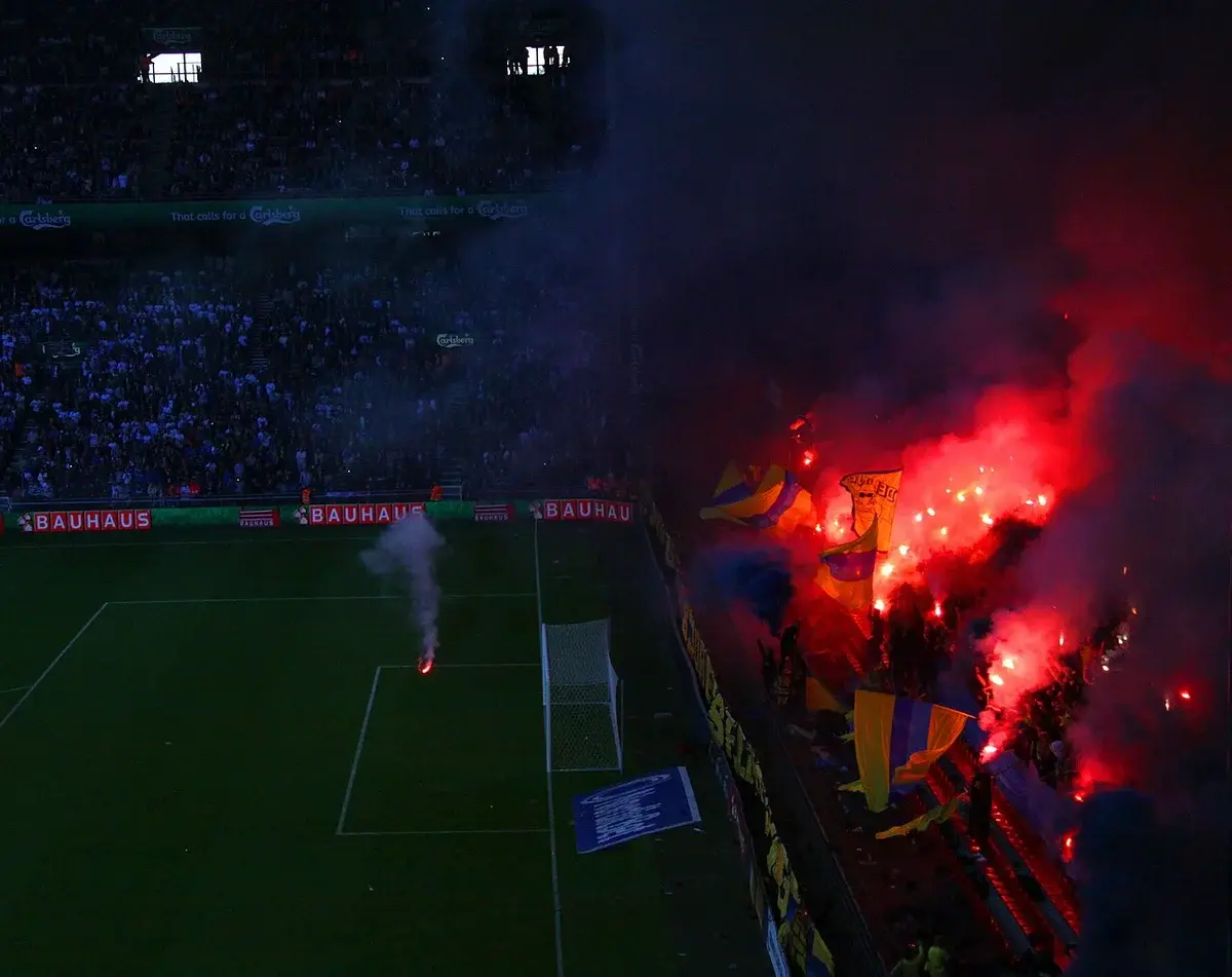 Football stadium scene with fans waving flags and red flares in the stands.