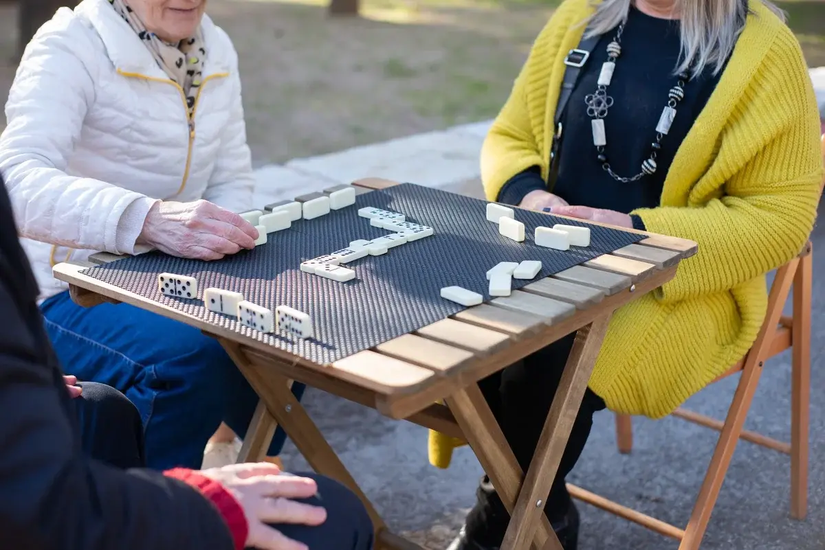 Older adults playing dominoes at an outdoor table.