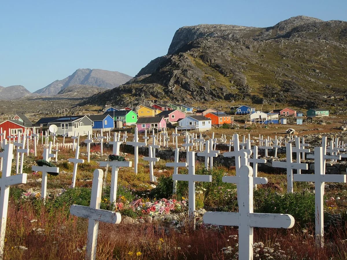 White crosses in a cemetery with colorful houses and a rocky hill in the background.