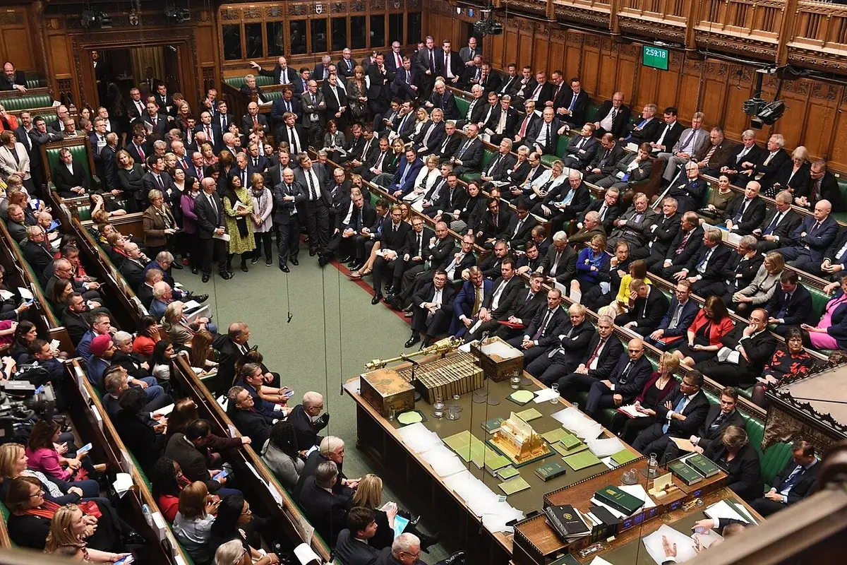 Wide view of the British House of Commons during a debate with MPs seated on green benches.