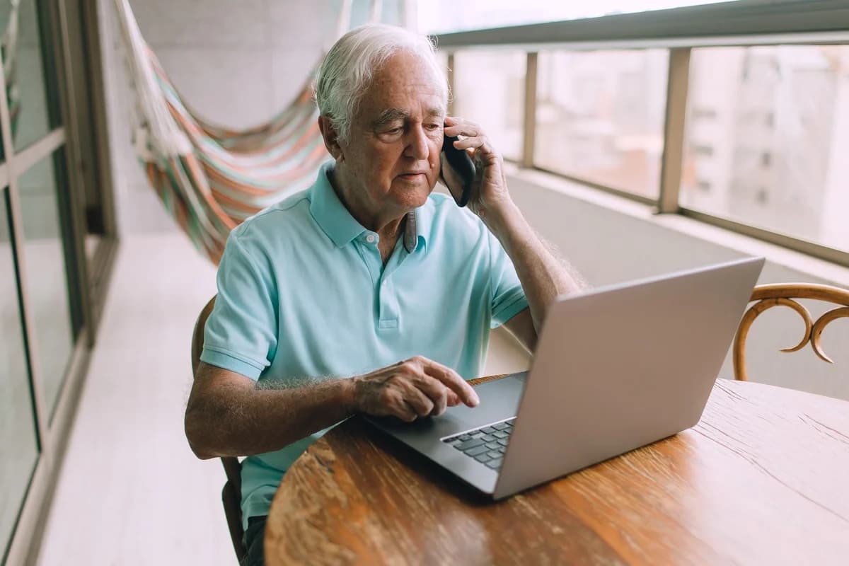 An older man uses a laptop while talking on a smartphone at home.