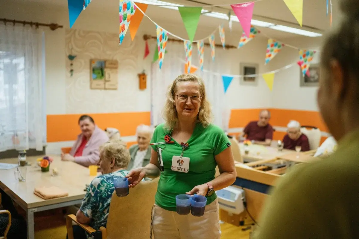 A caregiver serves drinks to elderly residents in a decorated care home room.