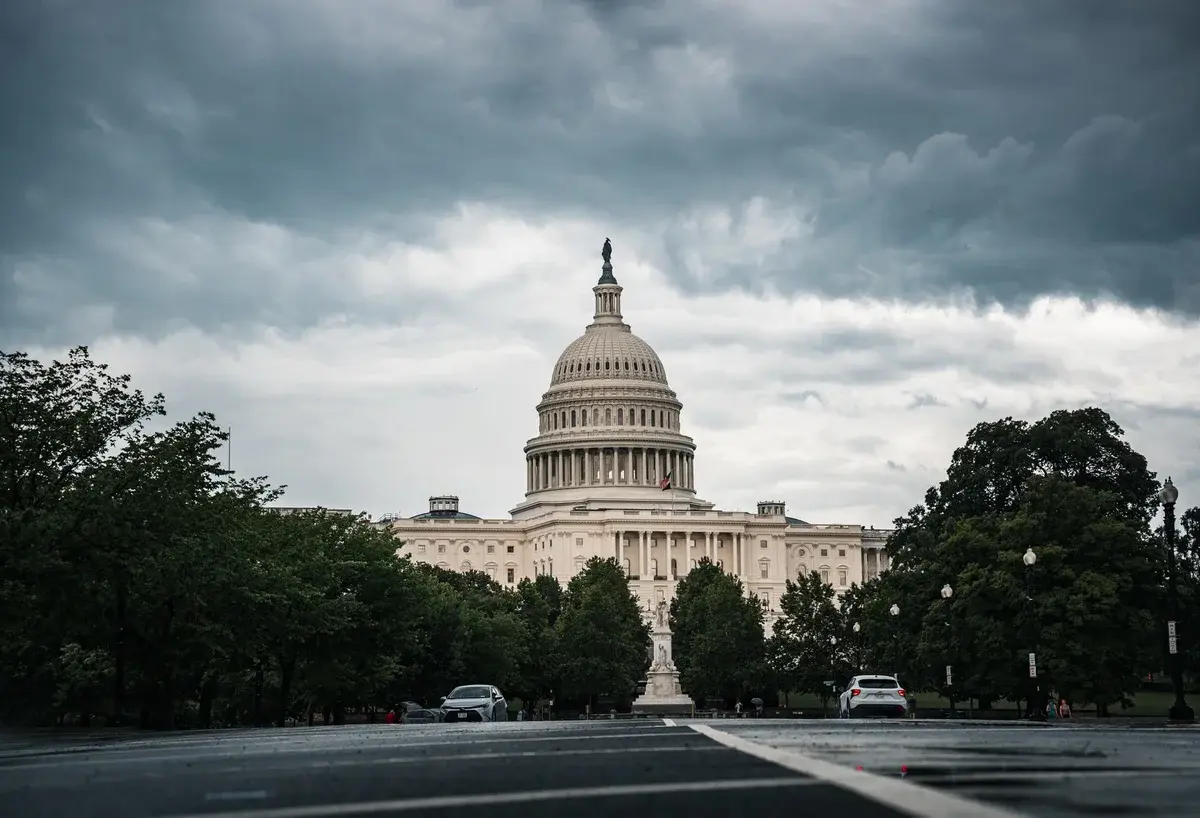 The US Capitol building under dark storm clouds with trees and cars in the foreground.