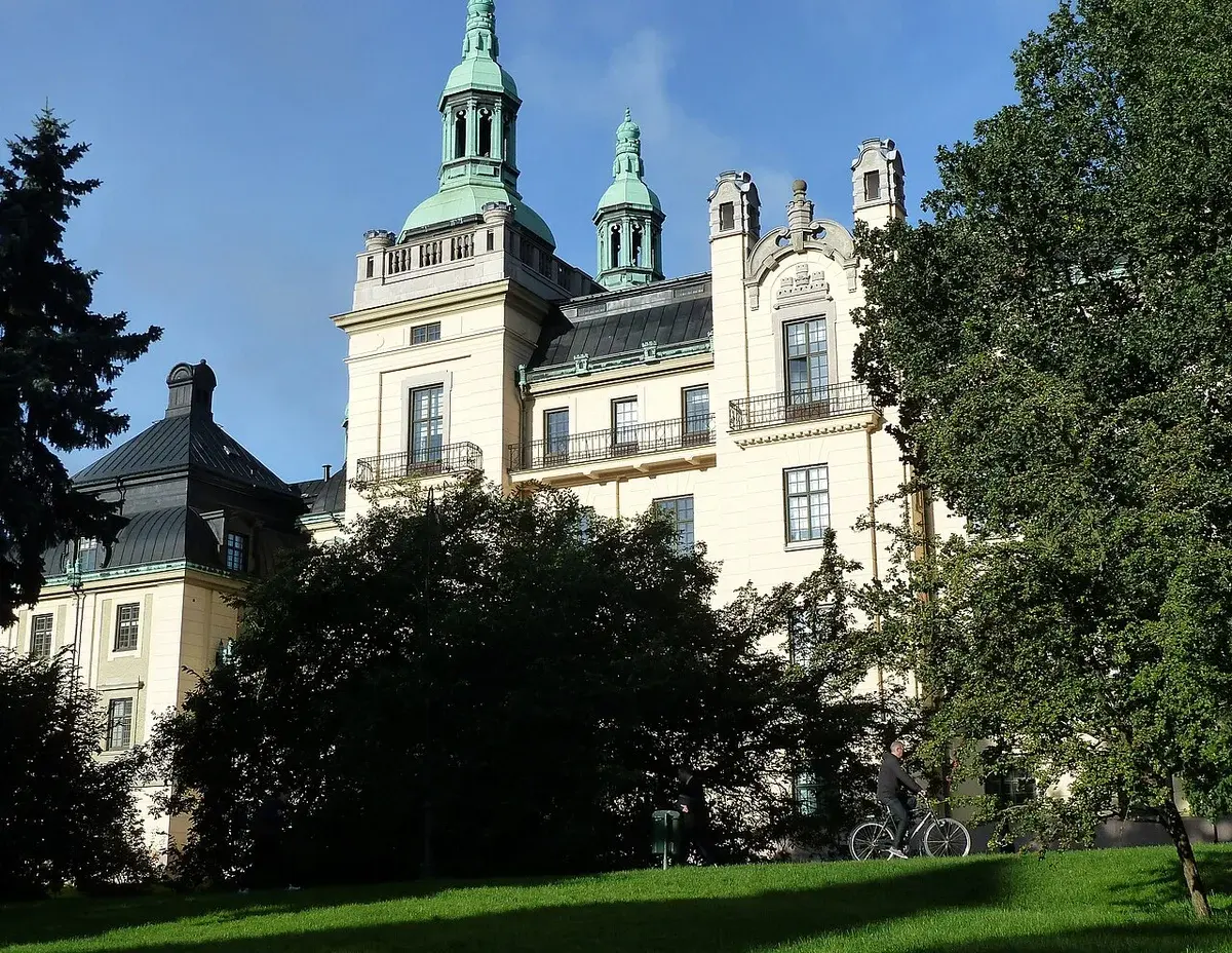 Historic building with green towers seen through trees in a park, with a cyclist in the foreground.