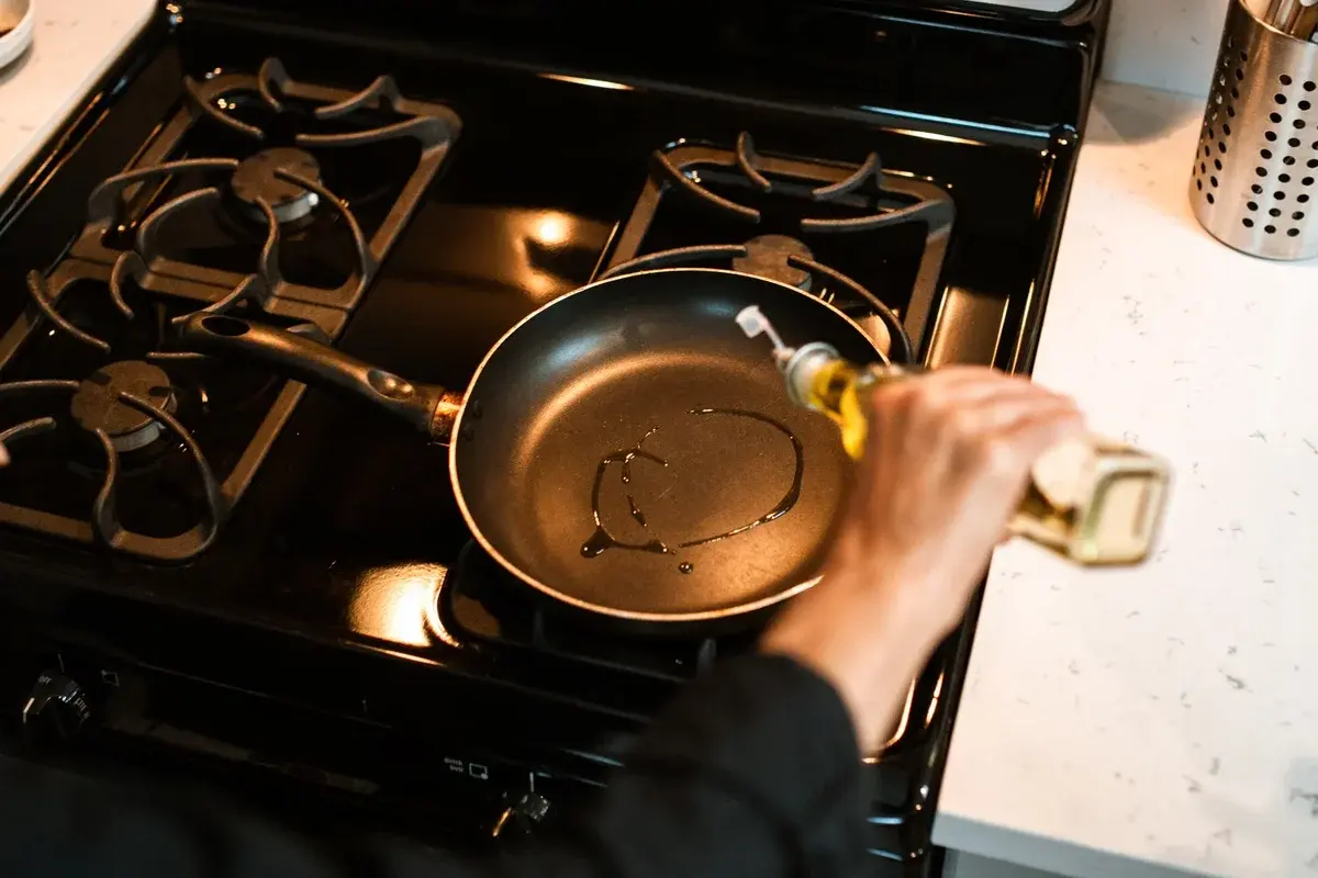 A person pours olive oil into a frying pan on a stove.