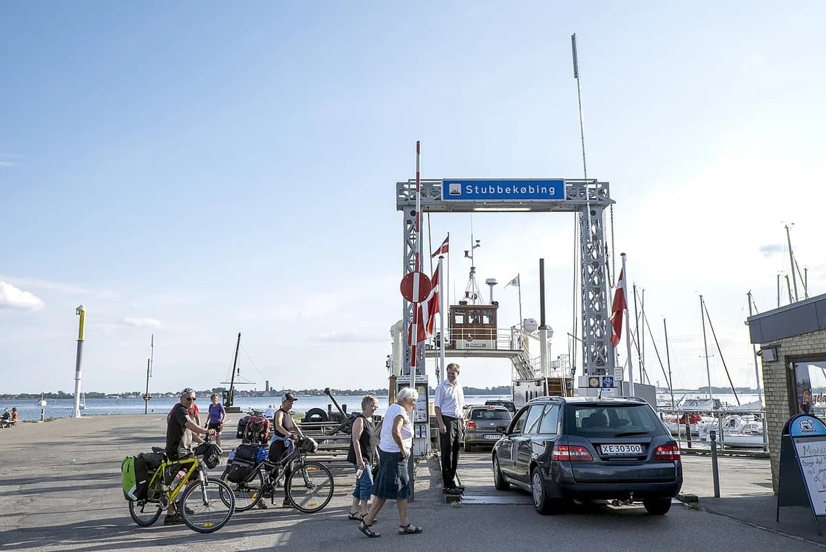 A small ferry at the Stubbekøbing terminal with cars, cyclists, and pedestrians nearby.