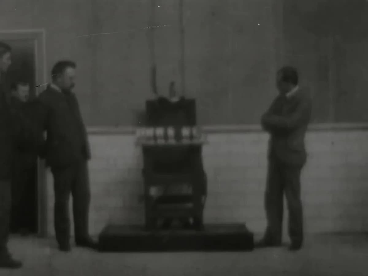 Black-and-white scene of men standing around an electric chair in a prison yard.