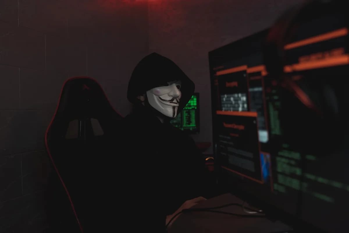 Masked hacker working at a computer in a dark room.