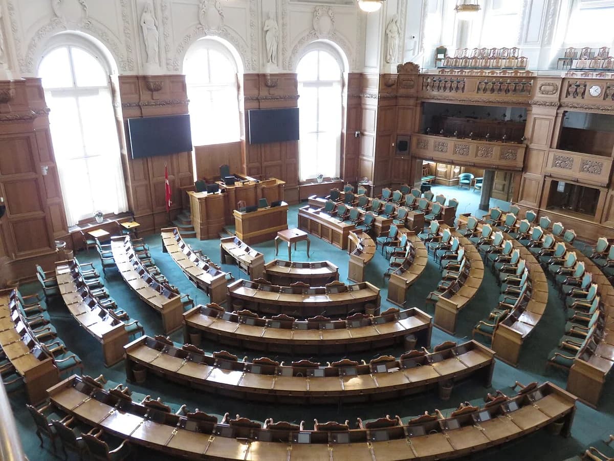 Empty Danish parliament chamber with curved rows of desks and green chairs inside Christiansborg Palace.