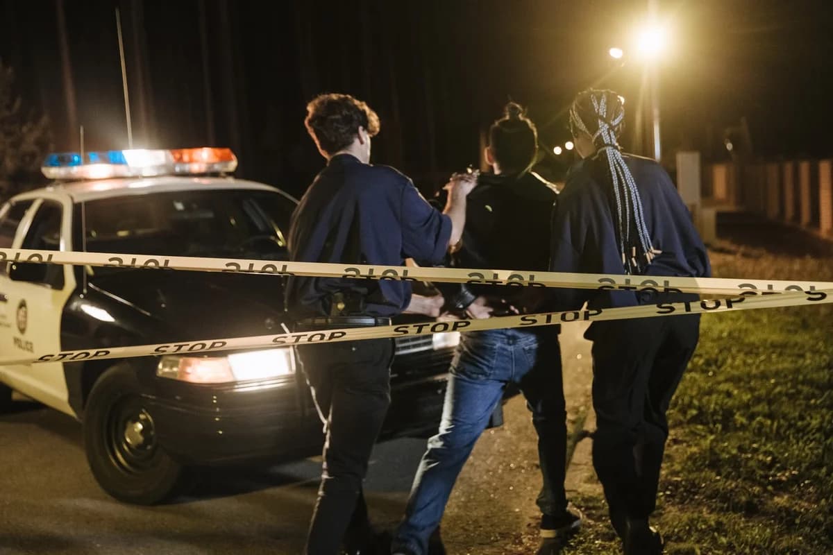 Police detain three people behind caution tape at night near a flashing patrol car.