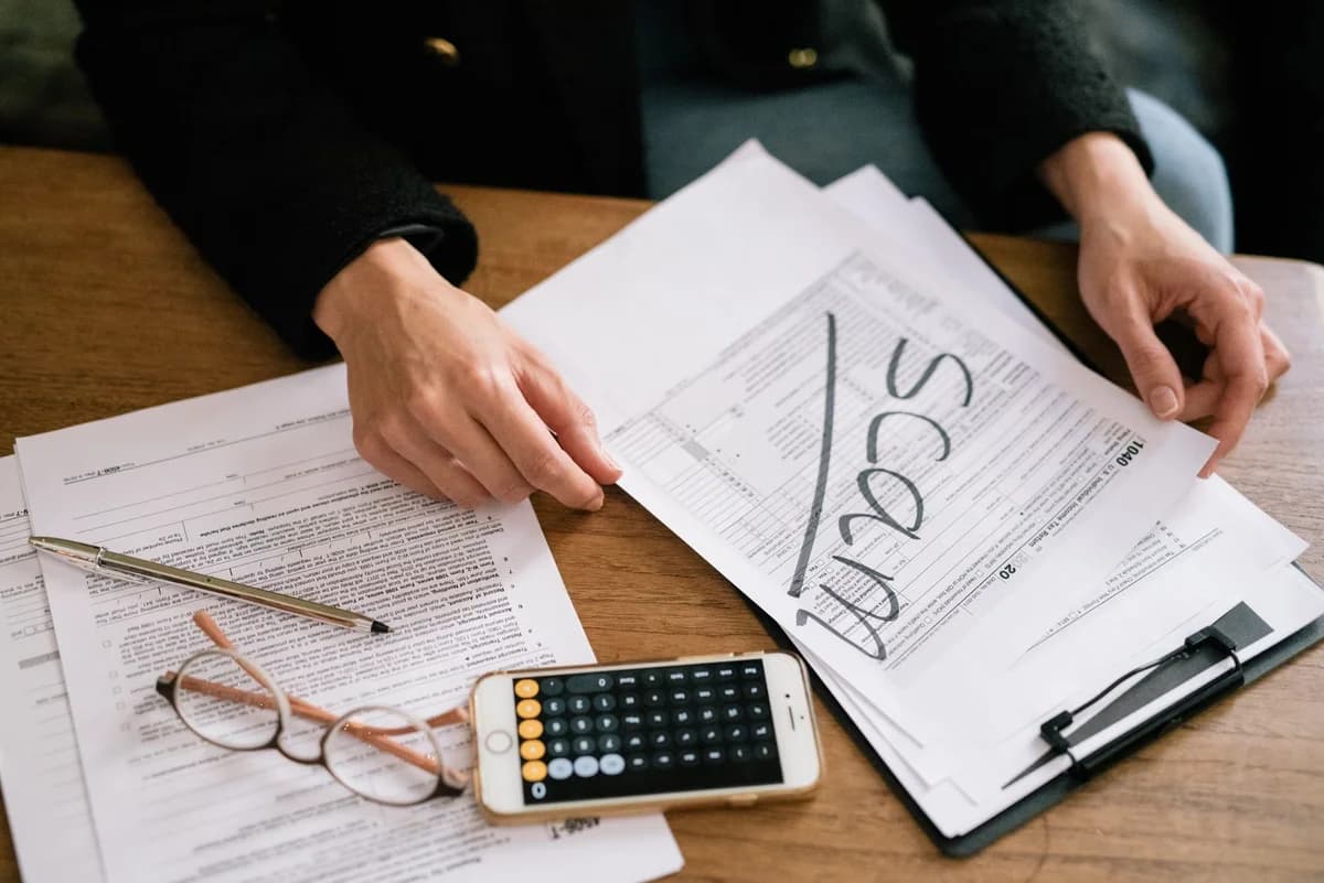 Hands reviewing tax papers marked "SCAM" beside a calculator phone, glasses, and documents on a desk.