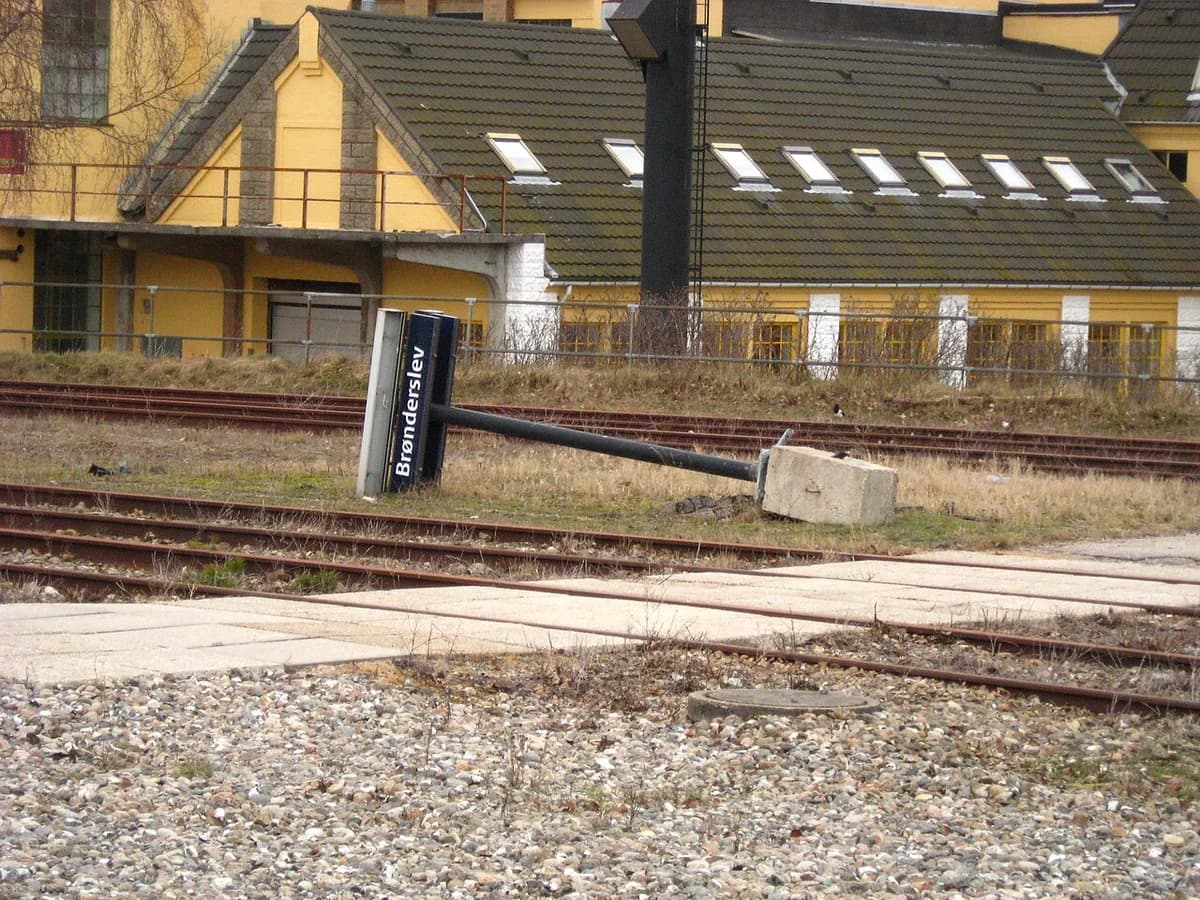 A fallen railway sign lies beside tracks near a yellow building.
