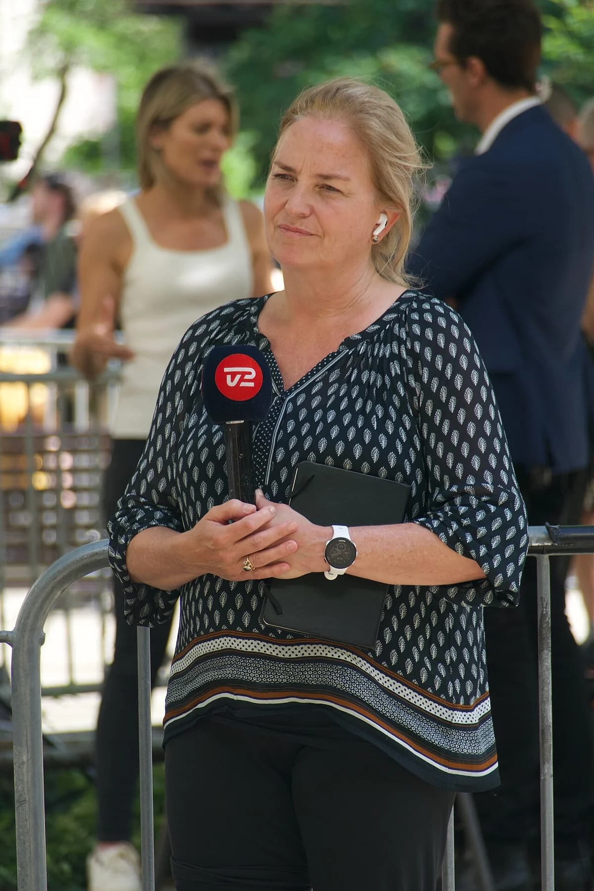 A TV2 reporter stands outdoors holding a microphone and tablet.