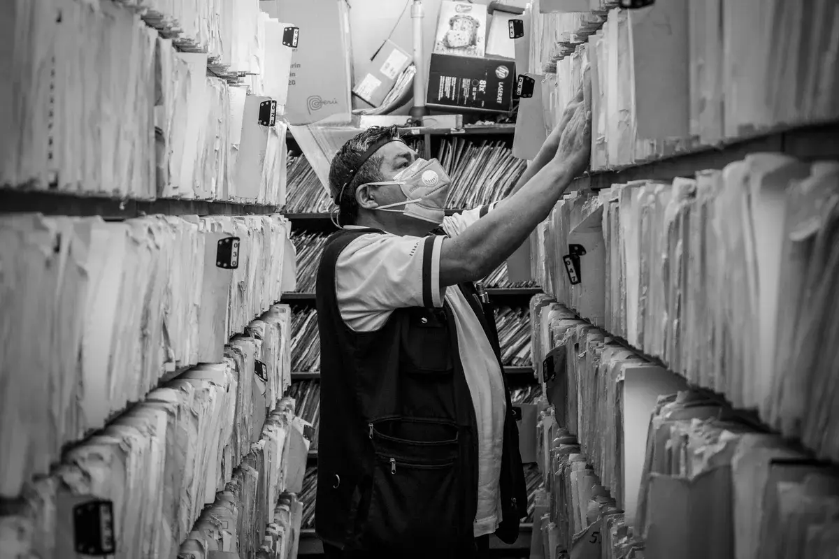 An older man wearing a face mask sorts files in a narrow archive aisle.