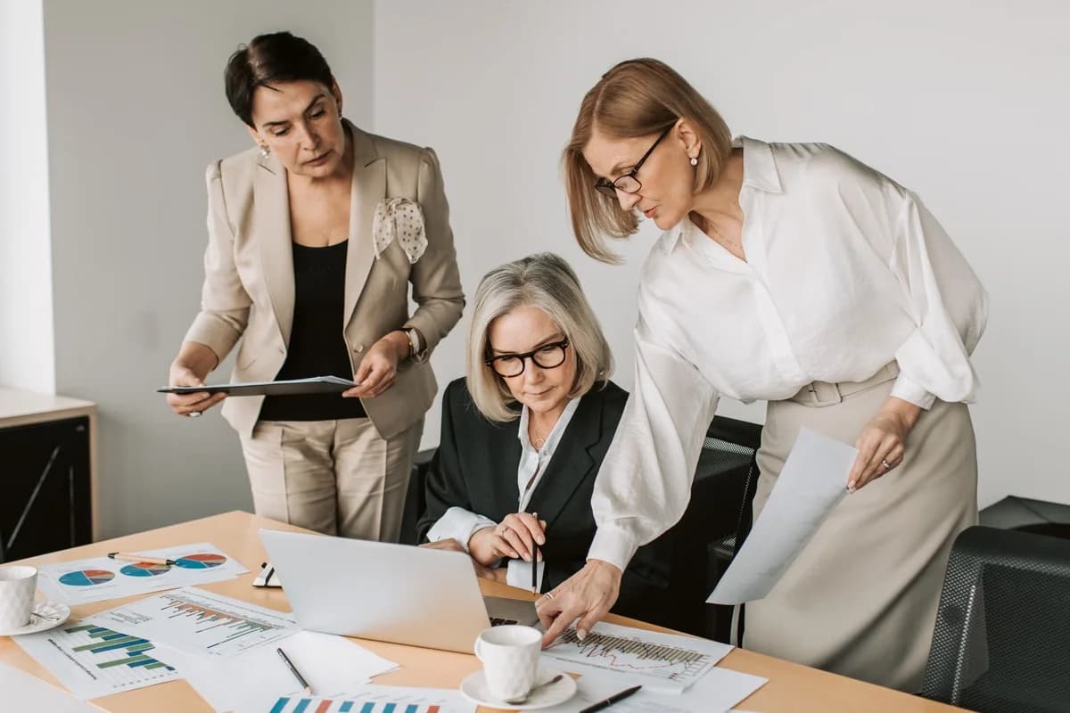 Three senior businesswomen reviewing charts and documents around a laptop in an office.