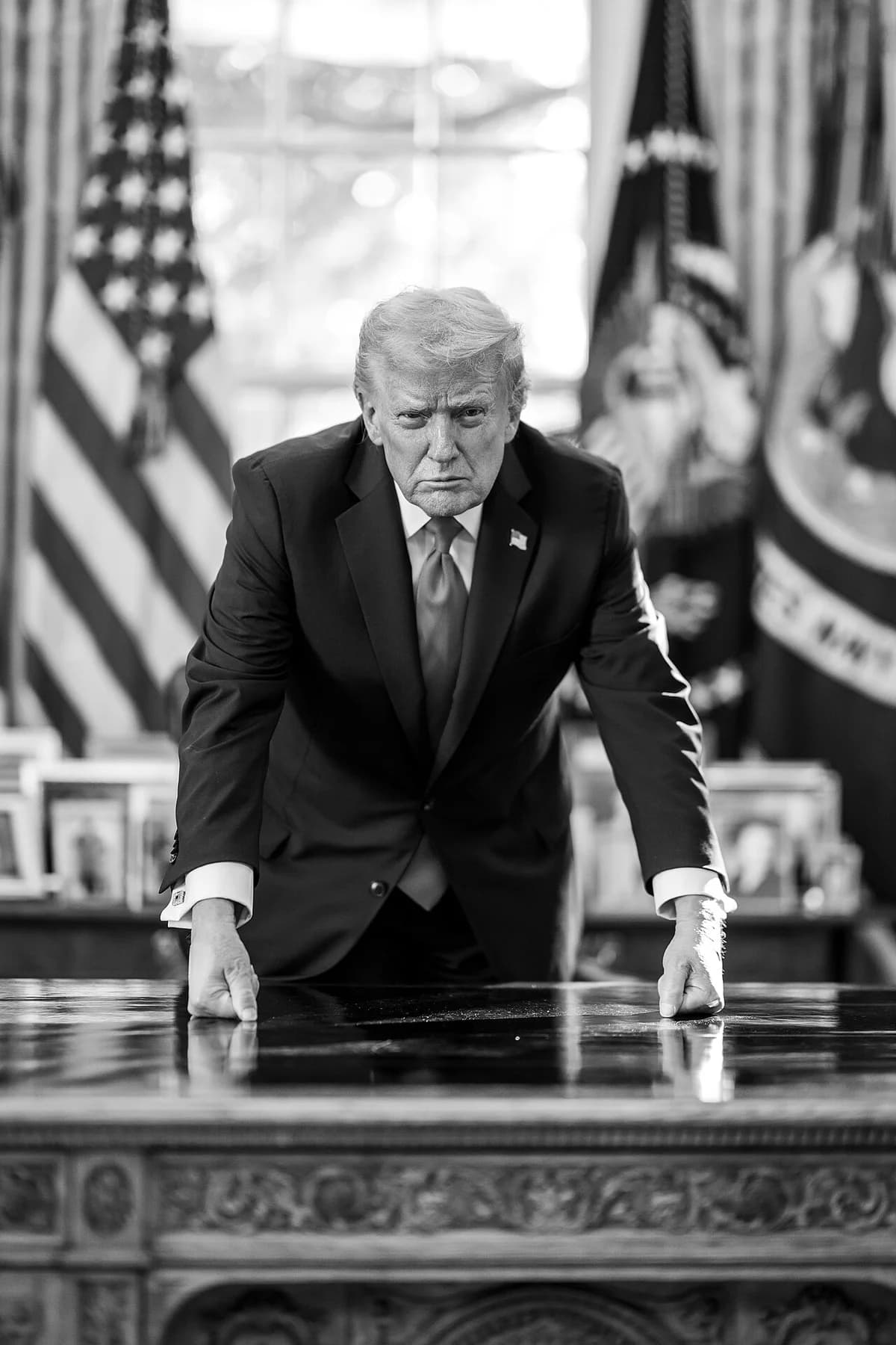 Black-and-white portrait of Donald Trump leaning on a desk with American flags behind him.