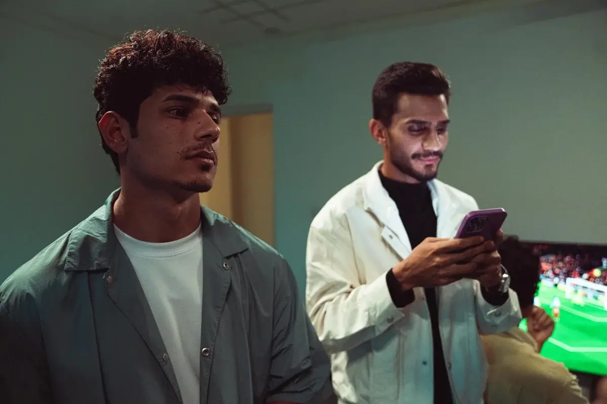 Two young men use smartphones in a room with a TV showing a sports game.
