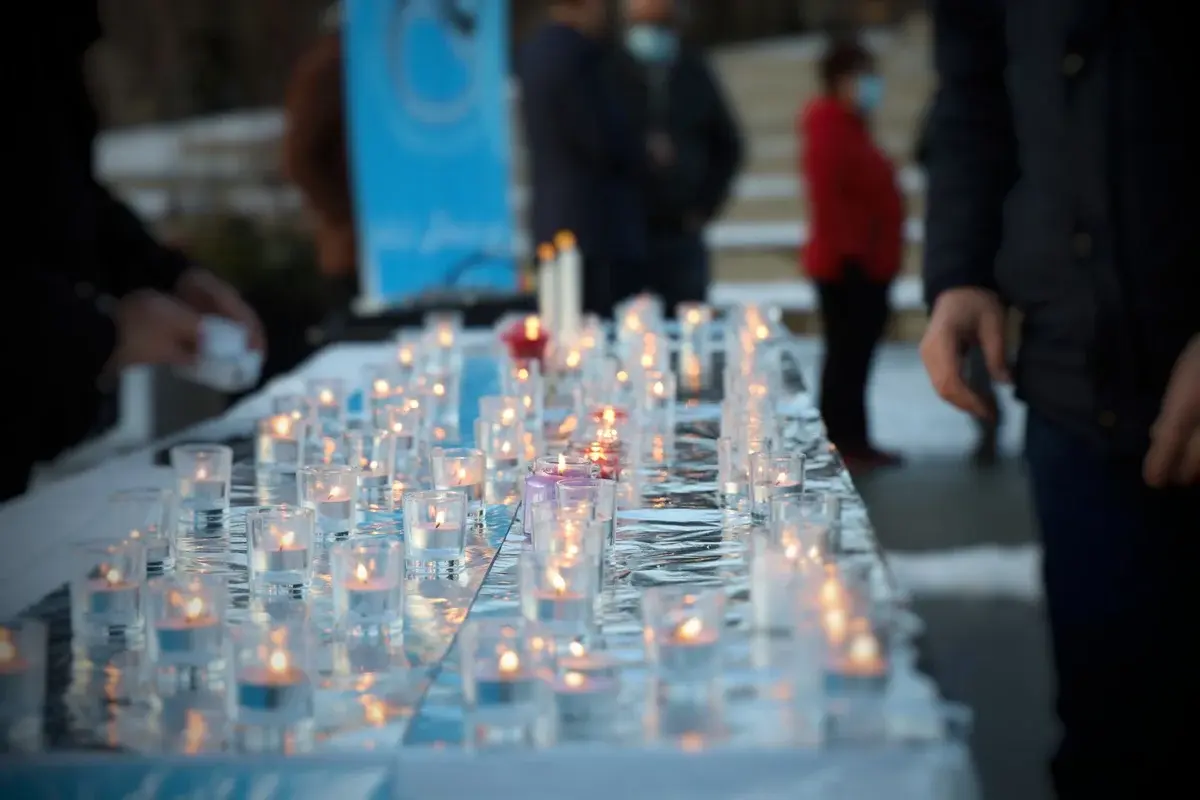 A table covered with many lit candles in glass holders, with people standing around it in the background.