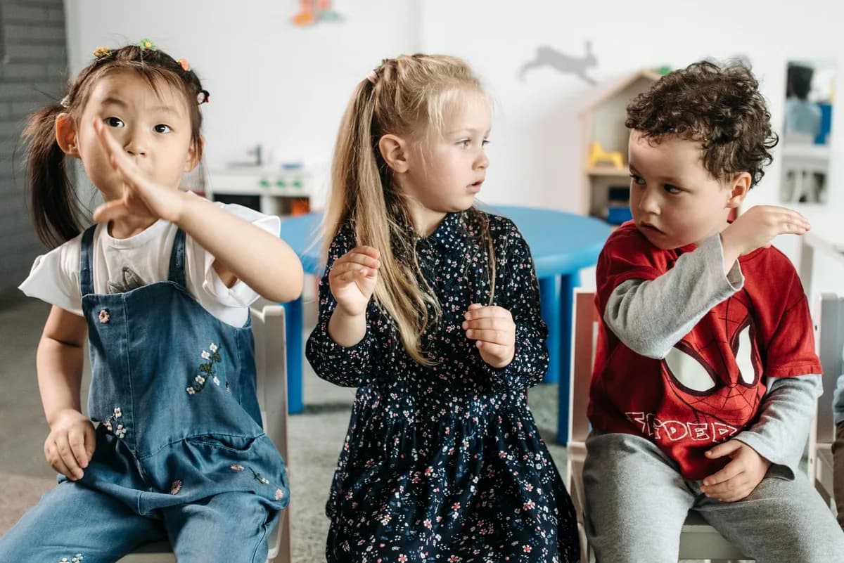 Three young children seated indoors, two making hand gestures.