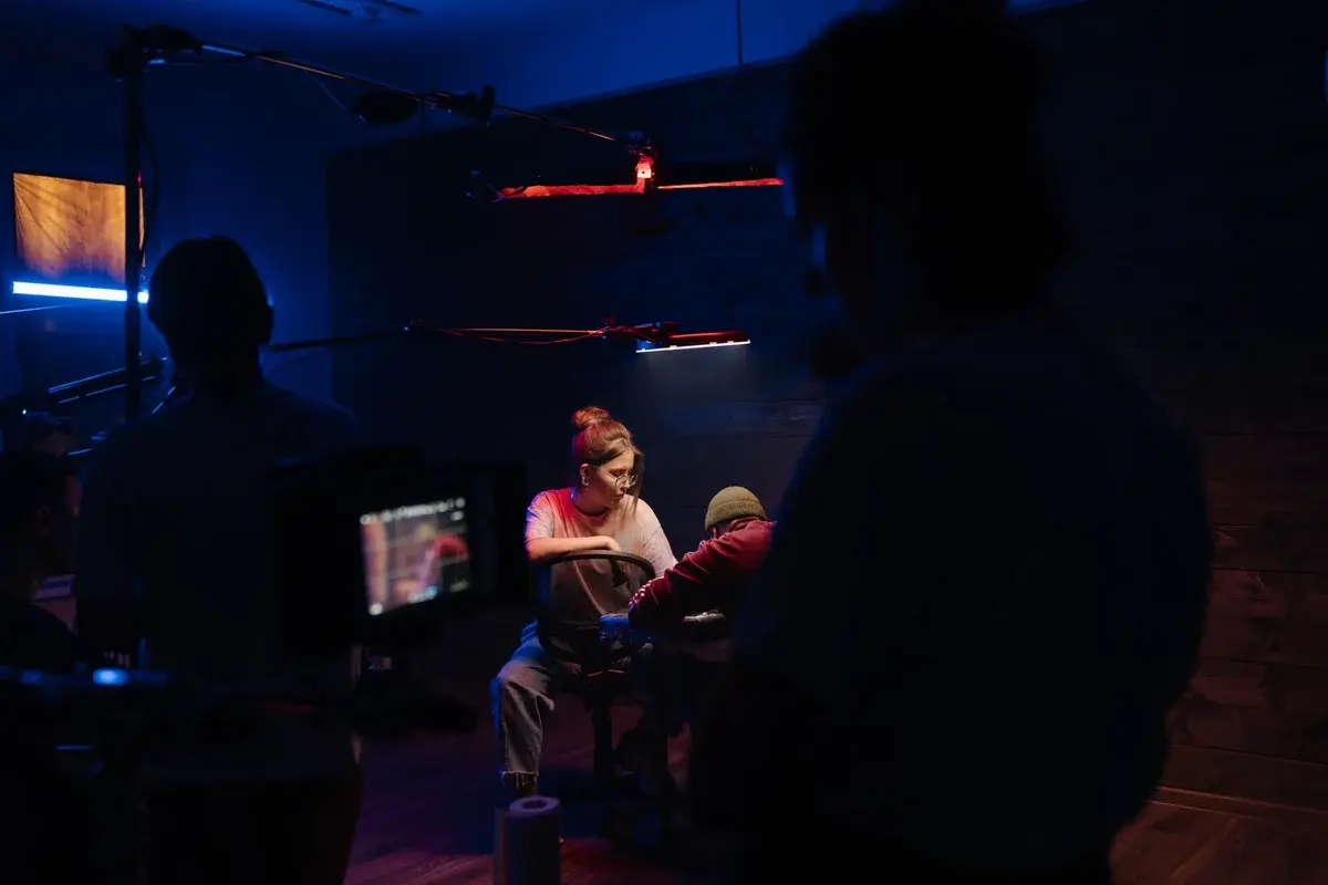 A neon-lit indoor film set with a camera crew filming two seated people.