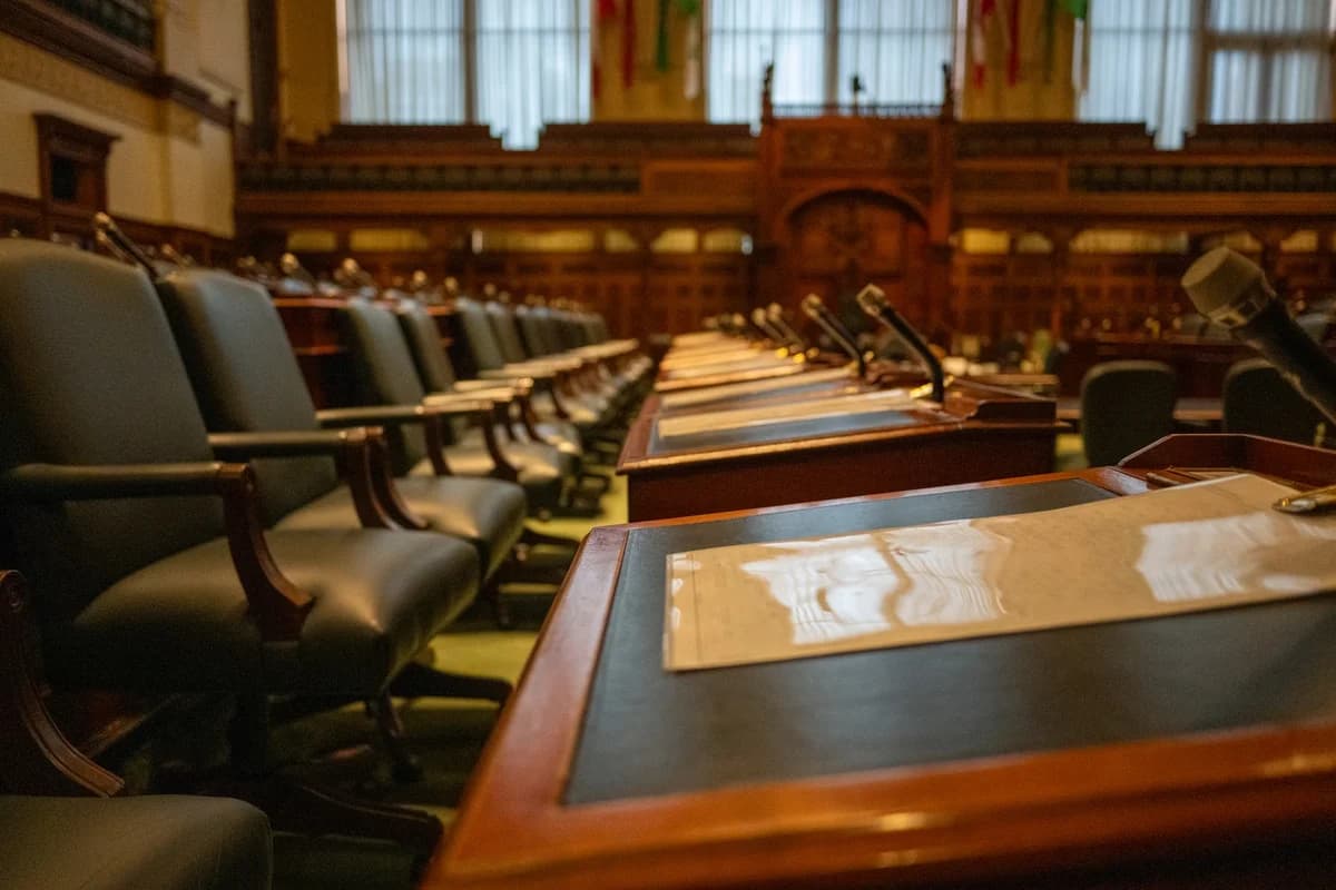 Empty legislative chamber with desks and microphones in a wood-paneled room.