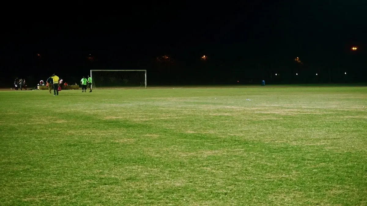 A lit soccer field at night with a goal and a few people near the left side.