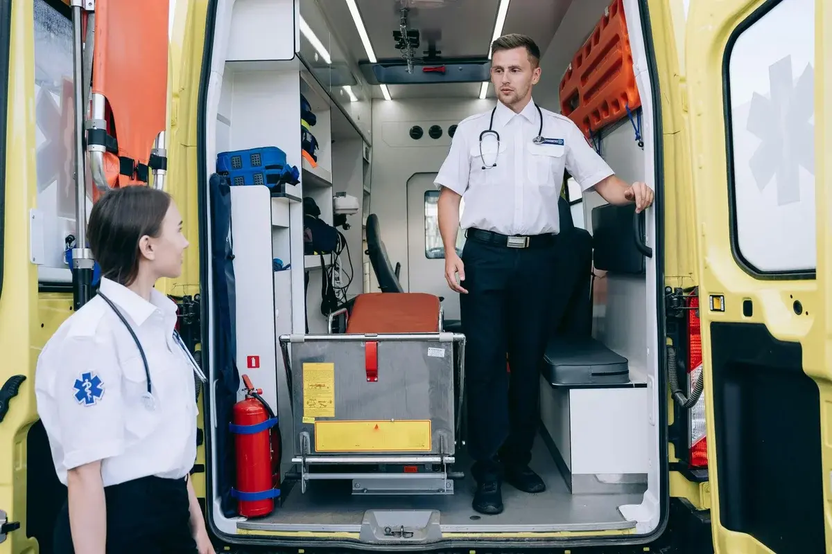 A male and female paramedic at the open rear of a yellow ambulance.