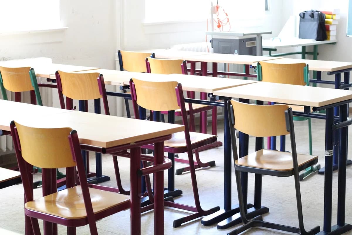 An empty classroom with wooden desks and chairs arranged in rows by large windows.
