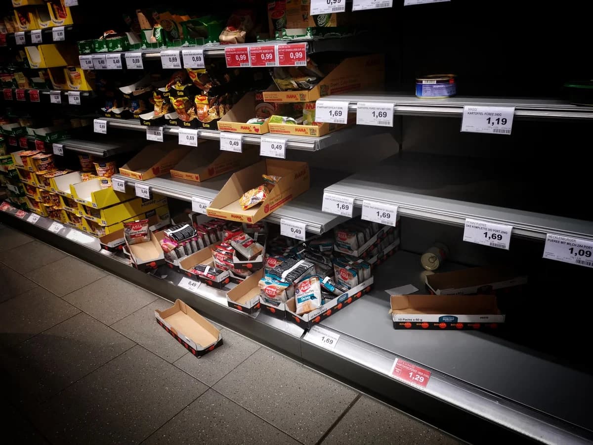 Supermarket shelves with a mix of products and empty spaces, plus price tags and cardboard boxes.