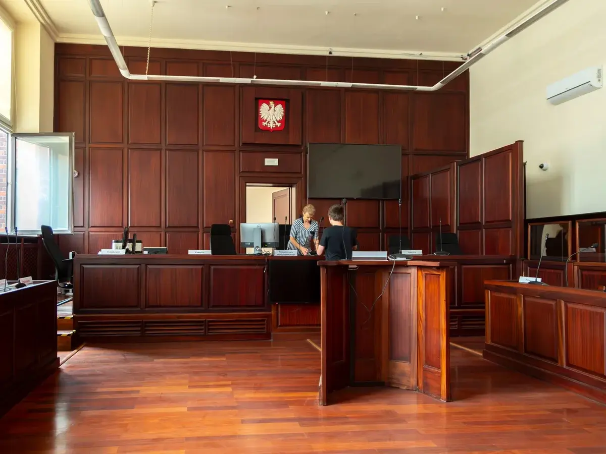 A bright Polish courtroom with dark wooden paneling, a Polish eagle emblem, and two people near the judge’s bench.