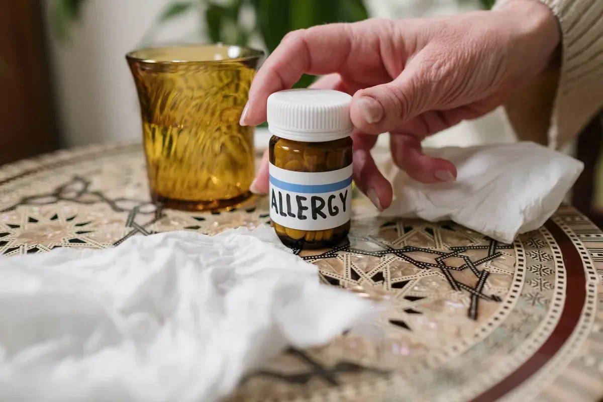 A bottle labeled allergy medicine beside tissues on a table, with a hand reaching in.