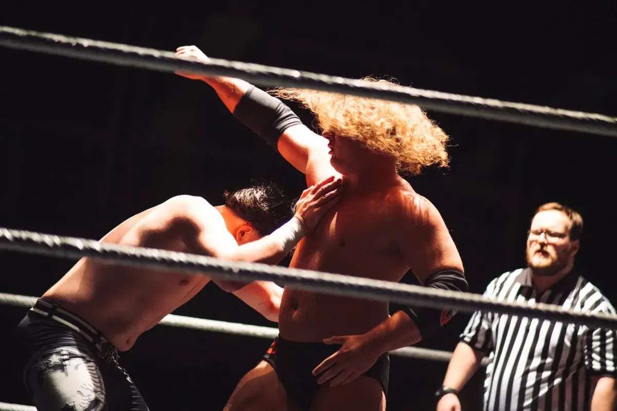 Two wrestlers grapple in a ring while a referee stands nearby.