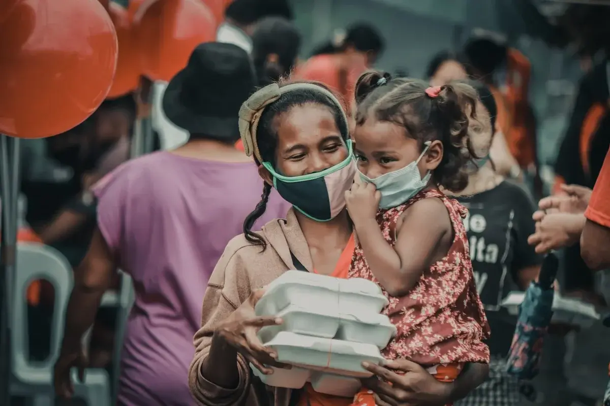 A masked mother carries stacked food packages while holding a masked child at an outdoor event with balloons.