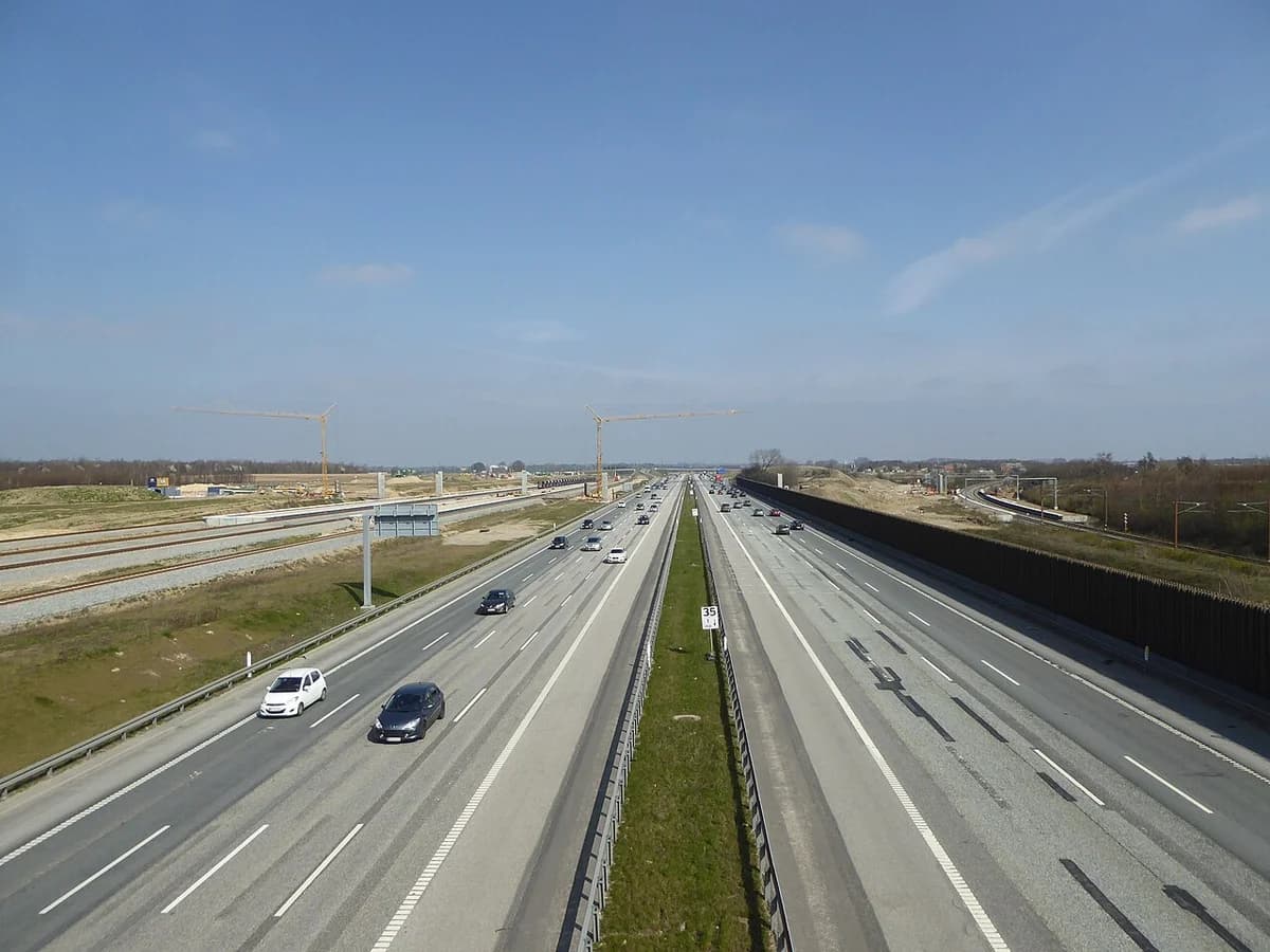 Cars travel along a wide motorway near construction work and cranes.