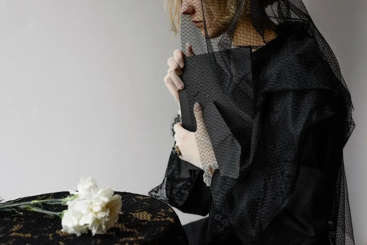 Woman in black mourning clothes holding a framed portrait with white flowers nearby