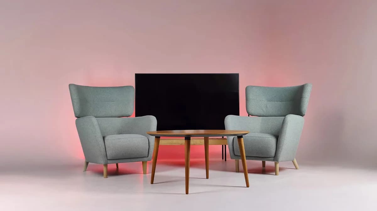 Two gray armchairs and a wooden table in front of a large TV screen with pink backlighting.