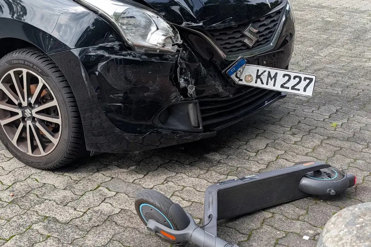 A black car with front-end damage and a fallen electric scooter on a paved street.