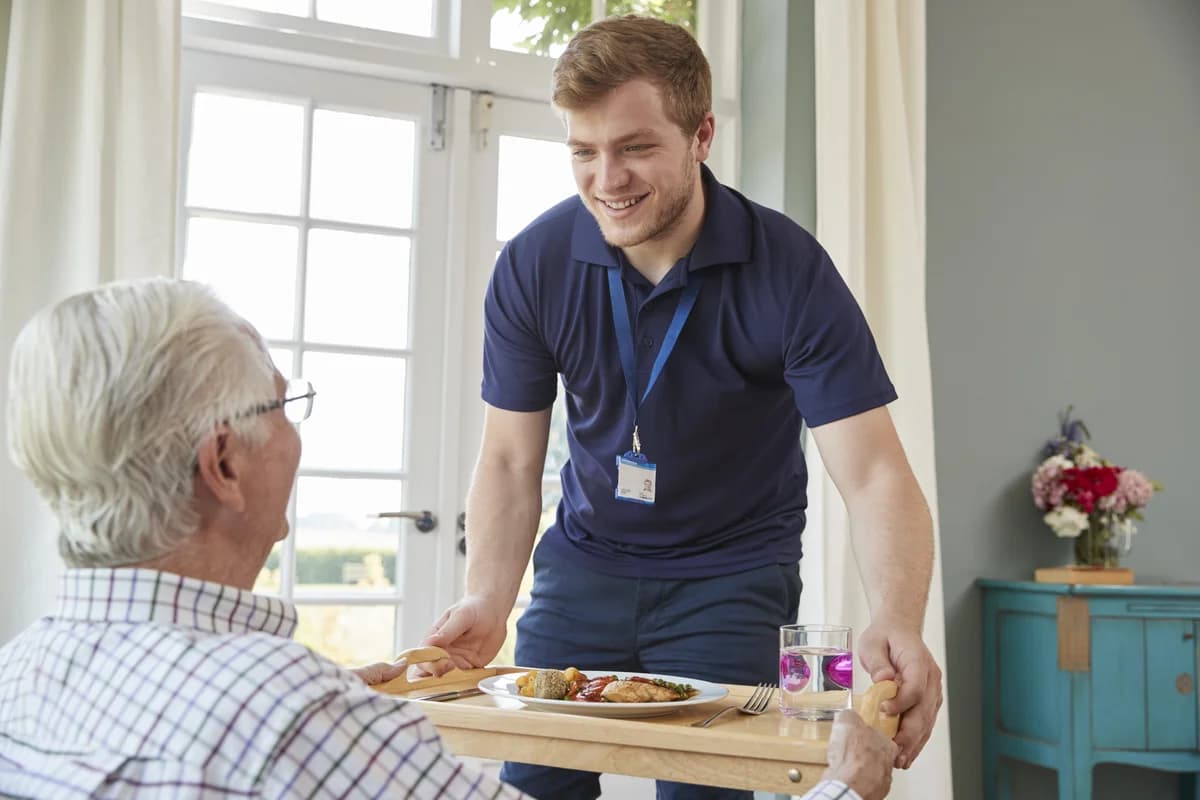 A caregiver serves dinner to an elderly man at home.