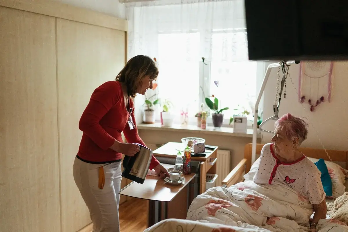 A care worker pours tea for an elderly woman sitting in bed.