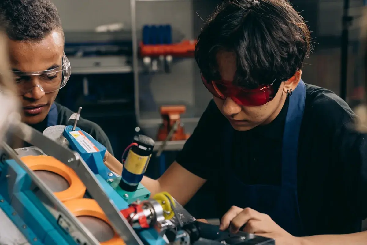 Two young people wearing safety glasses work on a device in a workshop.