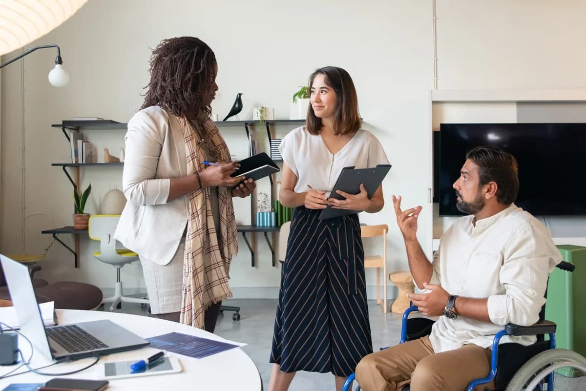 Three coworkers in an office, including a man in a wheelchair, discuss a project.