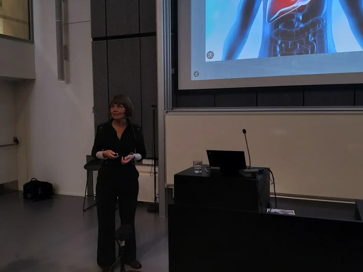 A woman speaks in a lecture hall beside a podium and projected screen.