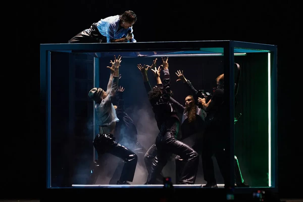 Singer on top of glass box with dancers inside reaching up