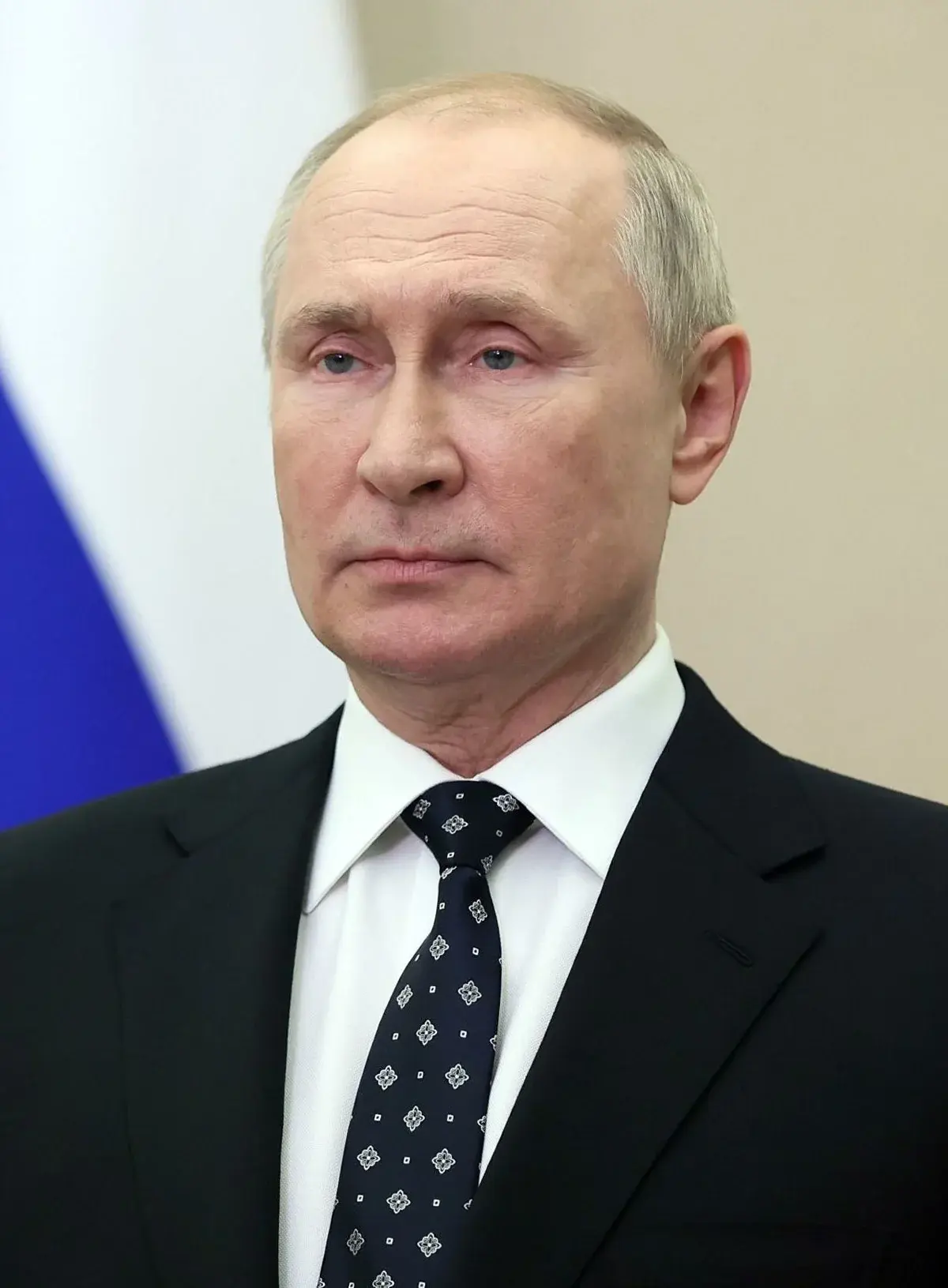 Close-up portrait of Vladimir Putin in a suit with a blurred Russian flag in the background.