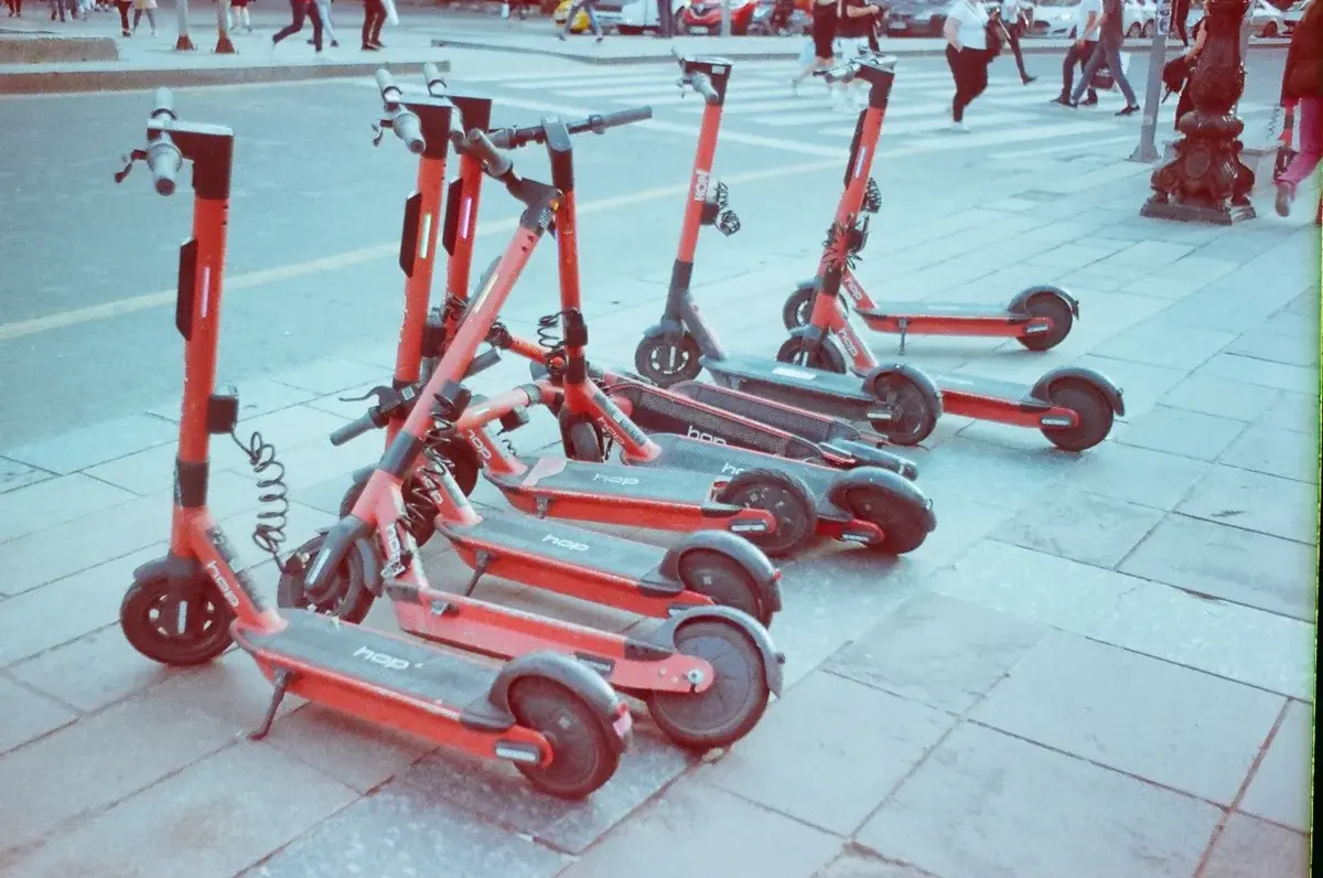 Red electric scooters lined up on a city sidewalk with pedestrians in the background