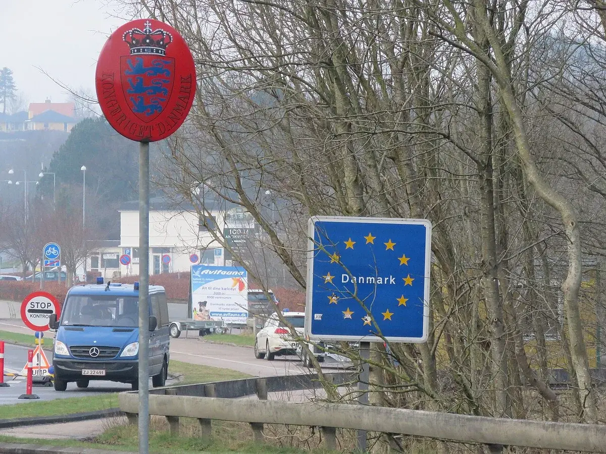 Border checkpoint with Denmark sign, stop signs, and vehicles on a roadway.