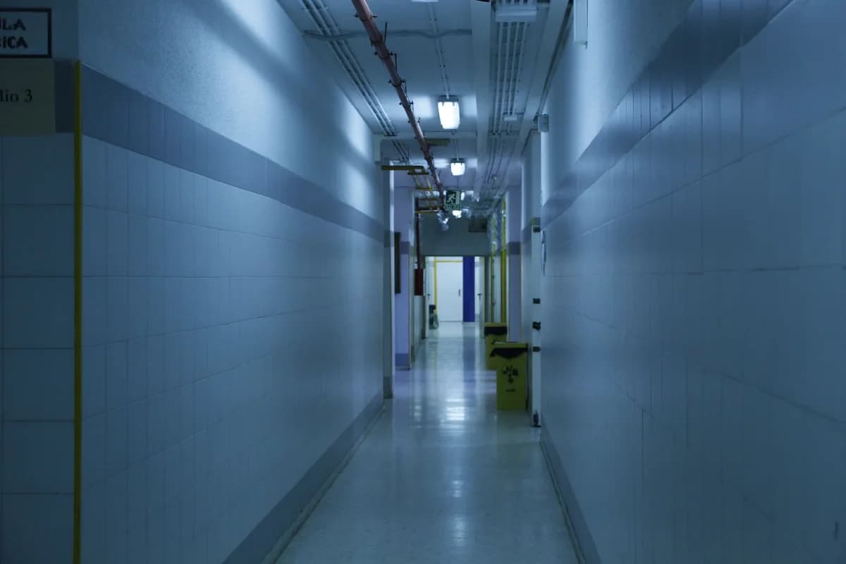 An empty, dimly lit hospital corridor with tiled walls and ceiling lights.